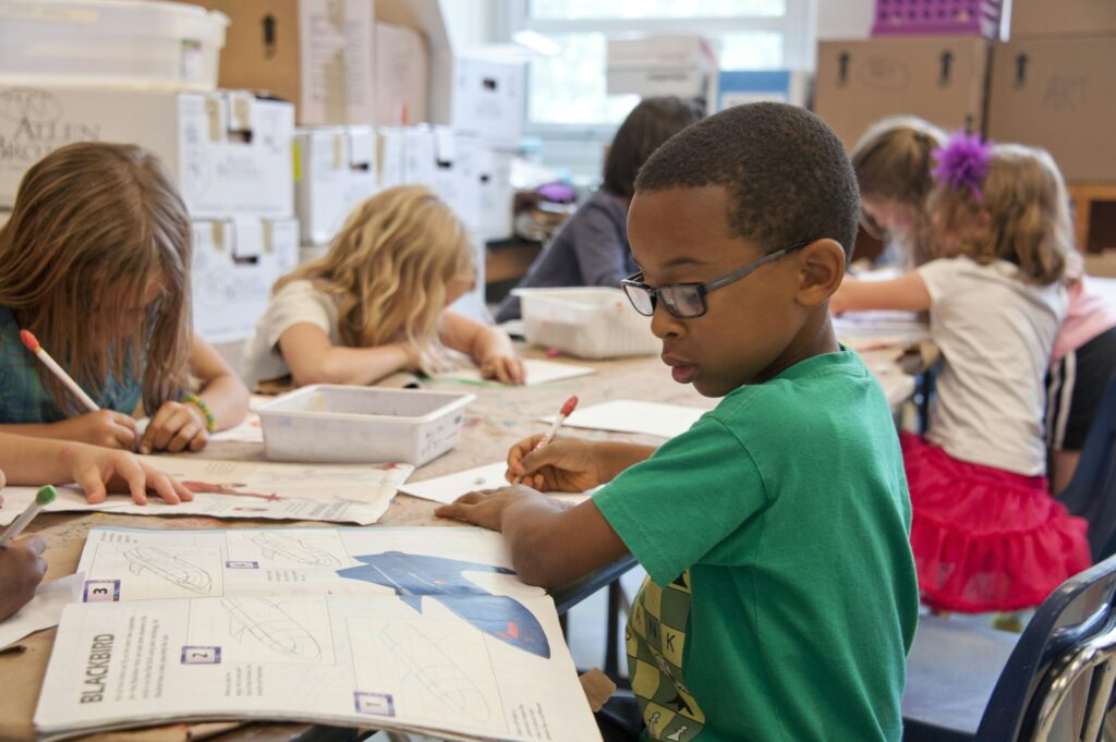 Primary school children writing in exercise books at a shared classroom table.