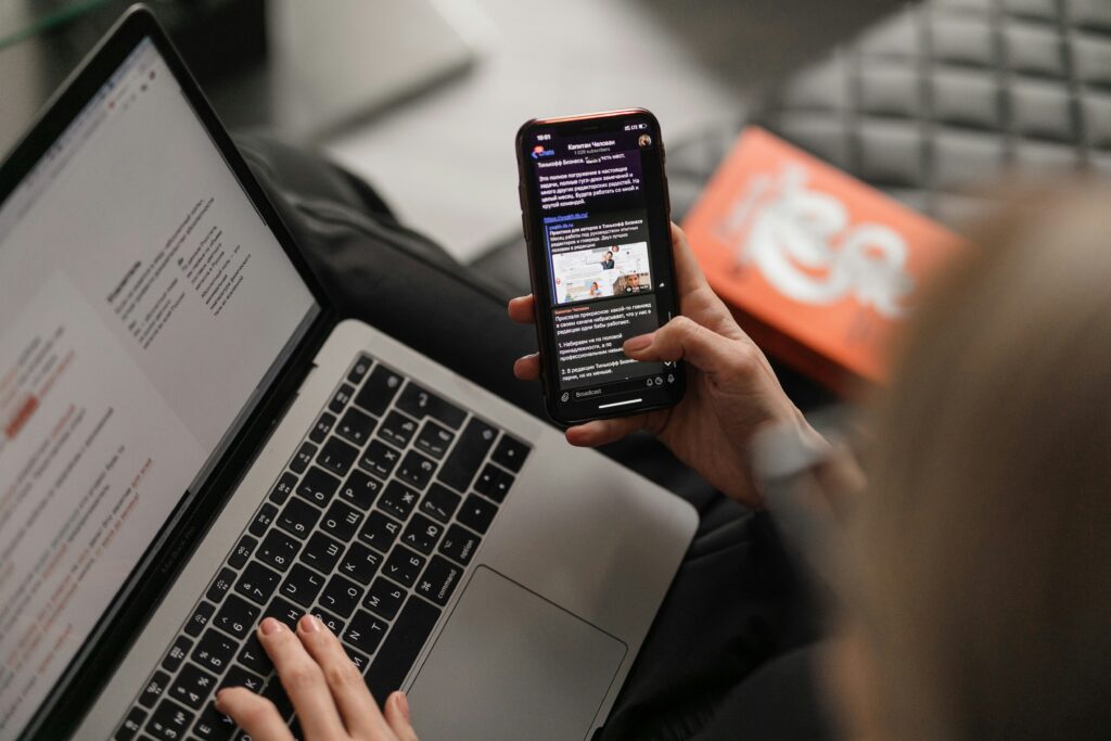 A person holding a phone displaying a news app while resting it on an open laptop.