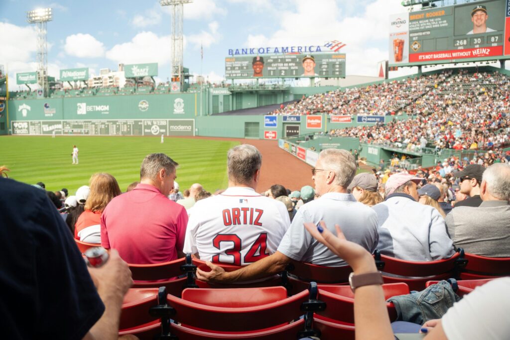 Fans in a packed baseball stadium watching a game, one wearing an Ortiz number 34 jersey, with the green outfield wall behind.