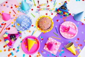 An overhead view of a birthday table with party hats, paper plates, cupcakes, and scattered confetti on a purple cloth.