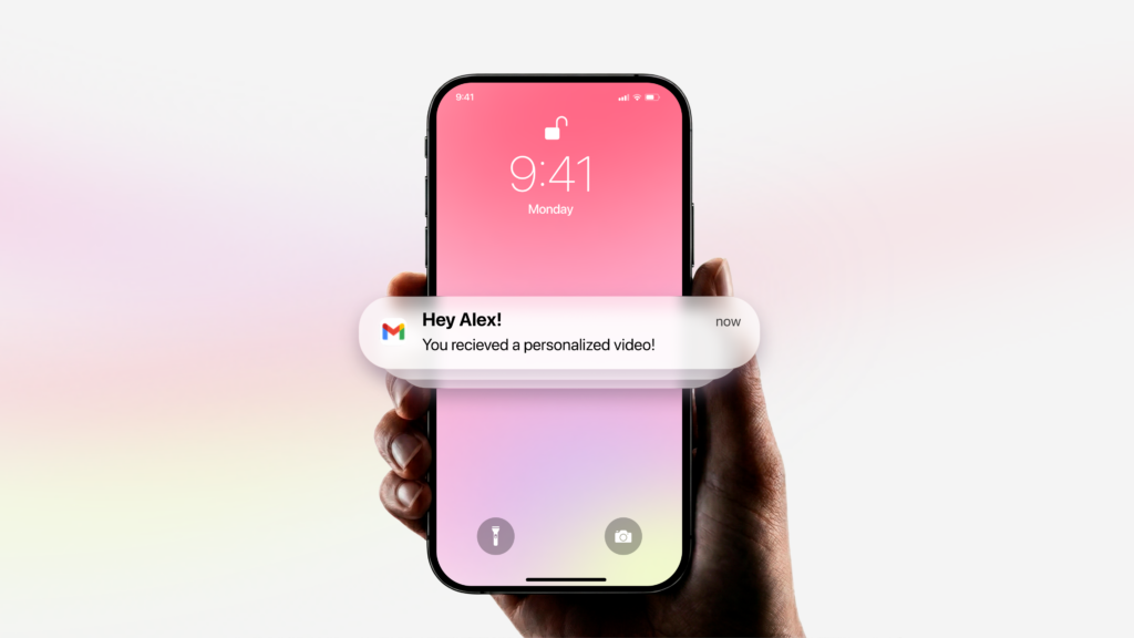 Hand holding a phone on a pink lock screen with an email notification reading Hey Alex, you received a personalized video.